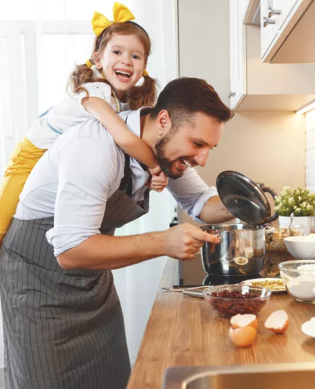 Vater mit Kind beim Kochen Ein Vater steht am Herd und kocht. Auf seinem Rücken sitzt ein junges Kind. Sie lachen beide.