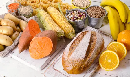 Auf einem weißen Tisch liegen Lebensmittel die viele Kohlenhydrate enthalten wie Brot, Süßkartoffeln, Kartoffeln, Mais, Orangen, Bananen, Nudeln, Getreide, Haferflocken