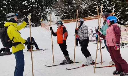 Kids und jugendliche Teilnehmer*innen des Skicamps an der Seite einer Skipiste, sie tragen Skikleidung und stehen auf jeweils einem Ski. Sie halten Stöcke in der Hand. Vor ihnen steht ein Skilehrer und erklärt, auch mit einem Stock in der Hand, etwas. 