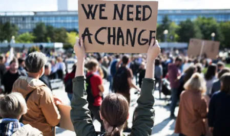 Menschengruppe bei einer Demonstration, im Vordergrund hält eine junge Frau ein Schild hoch "We need a change"