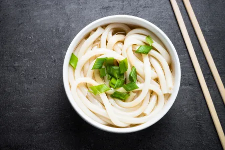 Udon Nudeln in Suppe in einer weißen Schüssel. Daneben liegen Essstäbchen