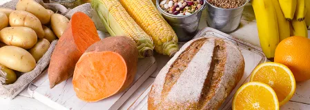Auf einem weißen Tisch liegen Lebensmittel die viele Kohlenhydrate enthalten wie Brot, Süßkartoffeln, Kartoffeln, Mais, Orangen, Bananen, Nudeln, Getreide, Haferflocken