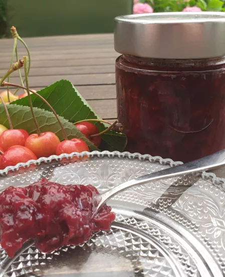 Kirsch-Vanille-Marmelade Die rote Kirschmarmelade steht in einem Glas auf dem Tisch. Ein Löffel liegt mit etwas Marmelade davor auf einem Teller, dahinter liegen ein paar Kirschen.