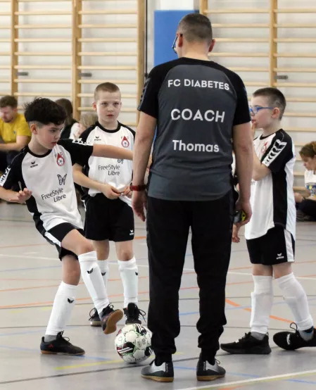 FC Diabetes Fußball Kinder spielen in einer Halle Fußball. Sie tragen schwarz-weiße Trikots. Im Vordergrund ist außerdem der Trainer zu sehen. Er trägt ein schwarzes Trikot mit der Aufschrift FC Diabetes Coach Thomas