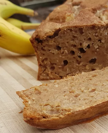 Bananenbrot mit Erdnüssen Bananenbrot mit Erdnüssen auf einem Holzbrett, mehrere Bananen im Hintergrund