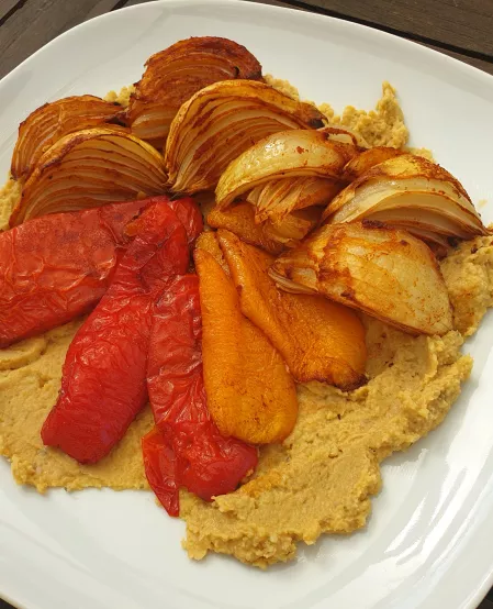 Ofengemüse mit Kichererbsenpüree Auf einem Bett von Kichererbsenpüree liegt im Ofen gegarte Paprika und Zwiebeln.