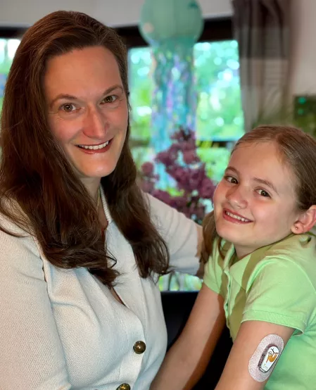 Mitgliederwahl Maren Sturny Maren Sturny sitzt seitlich und schaut in die Kamera. Rechts neben ihr sitzt ihre Tochter Sara-Léonie, die einen Sensor am Oberarm trägt. Beide lächeln in die Kamera.