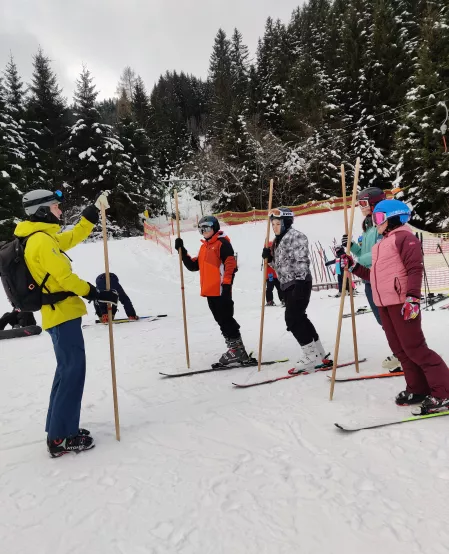 Skicamp Kids und jugendliche Teilnehmer*innen des Skicamps an der Seite einer Skipiste, sie tragen Skikleidung und stehen auf jeweils einem Ski. Sie halten Stöcke in der Hand. Vor ihnen steht ein Skilehrer und erklärt, auch mit einem Stock in der Hand, etwas.