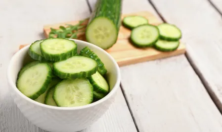 Gurke Gurkenscheiben in einer weißen Porzellanschüssel, im Hintergrund liegt eine angeschnittene Gurke auf einem Holzbrett