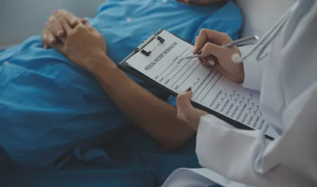 Krankenhaus Ein Patient liegt auf einem Krankenhausbett. Daneben eine Ärztin, die auf einem Klemmbrett Notizen macht.