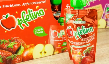 Ein einzelnes rotes Quetschie vor einer Packung Quetschies. Es hat einen grünen Deckel und ist die Sorte Apfel Erdbeere. 