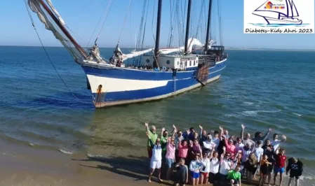 Eine Gruppe Kinder und Erwachsener am Strand vor einem Segelschiff. Sie recken die Arme in die Luft. Das Bild wurde bei Diabetes-Kids Ahoi 2023 aufgenommen.