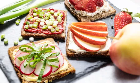 Auf einer dekorativen Platte sind vier Fitnessbrote angerichtet. Sie sind jeweils mit Frischkäse, Tomatenmark oder Senf bestrichen und mit Apfelschreiben, Radiesschen, Erdbeeren oder Frühlingszwiebeln belegt. 