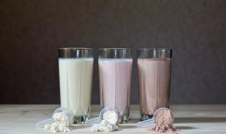 Formula-Nahrung Drei große Gläser mit Shakes. Davor liegen jeweils Löffel mit dem dazugehörigen Pulver. Der erste Shake sieht aus wie ein Vanille-Shake. Der zweite wie Beere und der dritte wie Schokolade.