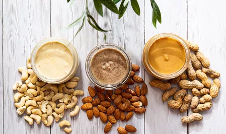 Nussmus Auf einem Tisch stehen drei Gläser mit Nussmus, vor ihnen liegen die jeweiligen Nüsse: Cashews, Mandeln und Erdnüsse