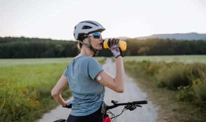 Eine Radfahrerin mit Helm und Sportbrille auf einem Feldweg. Sie schaut nach rechts während sie trinkt. An ihrem Arm ist ein CGM-Gerät zu sehen