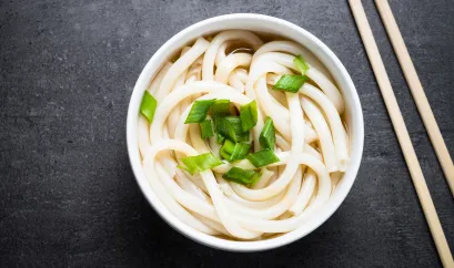 Udon Nudeln in Suppe in einer weißen Schüssel. Daneben liegen Essstäbchen