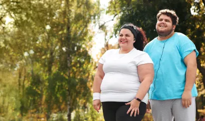 Eine Frau und ein Mann mit Adipositas in einem Park. Sie tragen Sportkleidung und lächeln.