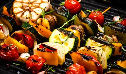 Gemüsespieße auf einem Grill. Tomaten, Zucchini, Paprika, Ziebel und Pilze sind auf dem Spieß zu finden. Auf dem Grill liegt außerdem eine Knoblauchknolle.