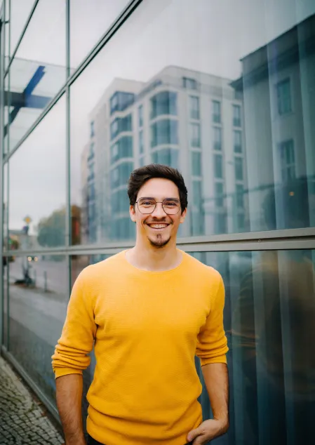 Ivo Rettig Ivo Rettig hat dunkle Haare, eine dunkle rechteckige Brille und trägt einen orangenen Pulli. Er lacht in die Kamera. Im Hintergrund spiegeln sich Hochhäuser.
