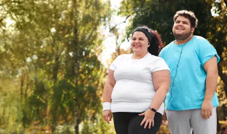 Eine Frau und ein Mann mit Adipositas in einem Park. Sie tragen Sportkleidung und lächeln.