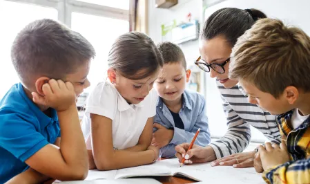 Vier Kinder an einem Tisch mit einer Lehrerin, die etwas erkl&auml;rt